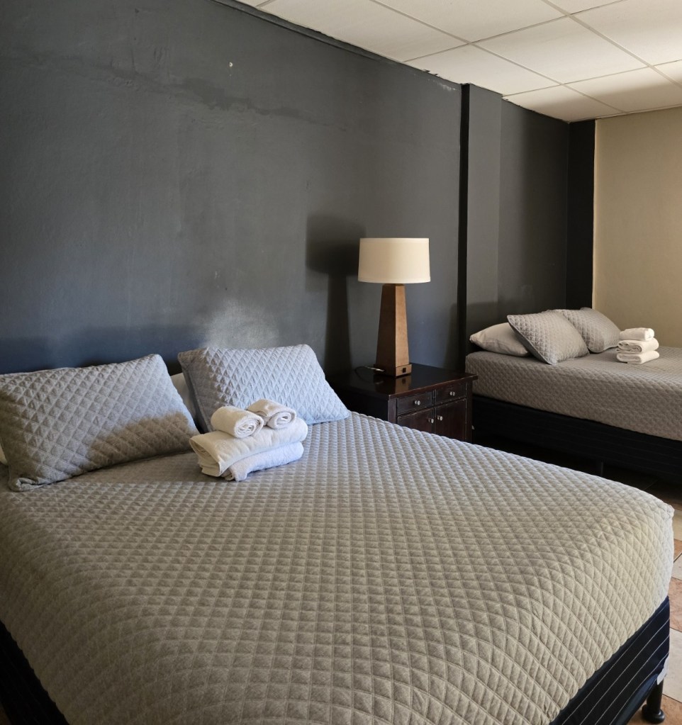 Habitación familiar con aire acondicionado con dos camas queen, lámpara de noche, toallas dobladas sobre la cama y pared gris